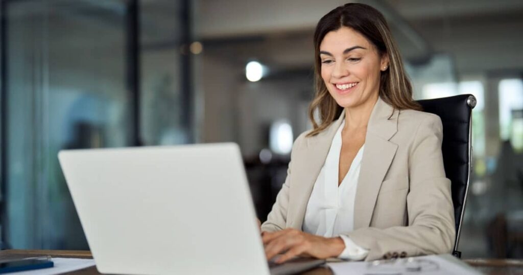 Une femme souriante assise à un bureau regarde l'écran de son ordinateur portable. 