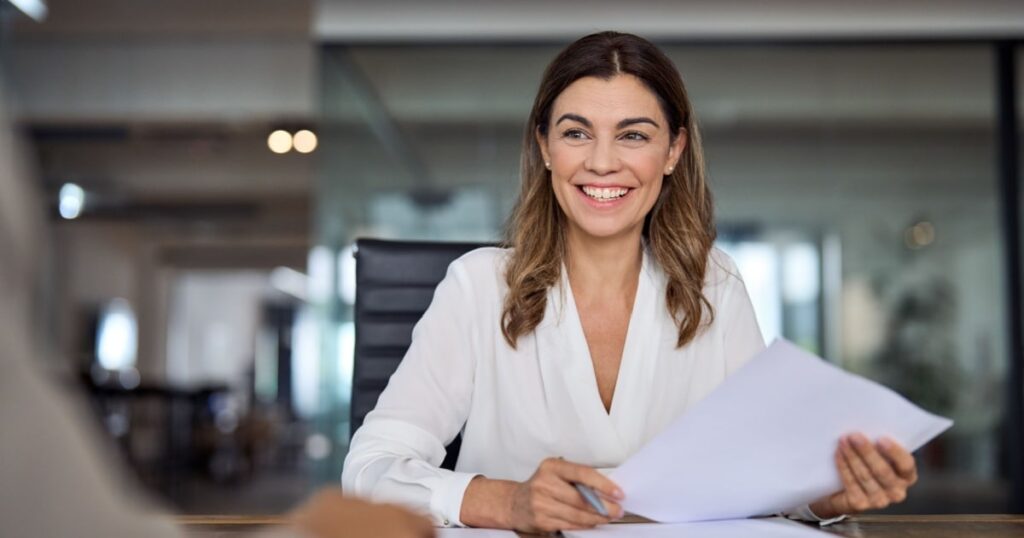 Une femme assise à une table de réunion sourit à son interlocuteur en tenant un document. 