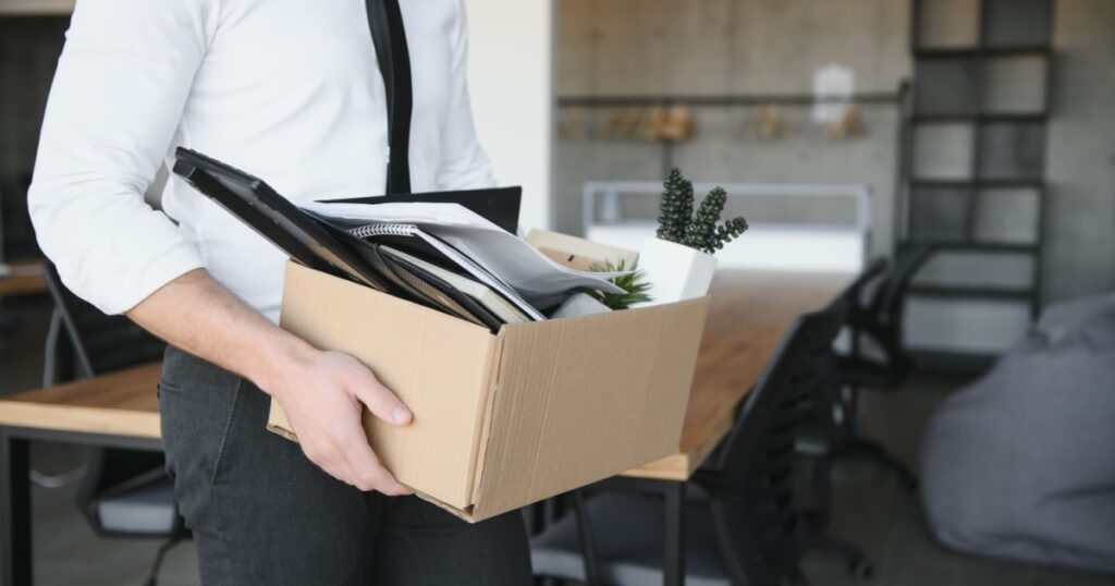 Un homme se tient debout dans un bureau, il tient un carton avec des affaires de bureau. 